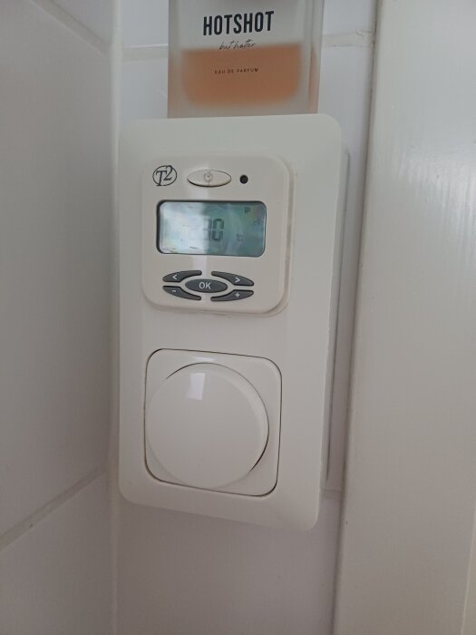 Vit väggmonterad termostat med display och knappar, bredvid en parfymflaska märkt "Hotshot".