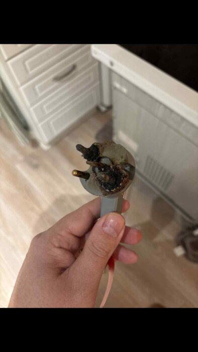Bränt och deformerat spisuttag med metallstift, hållet i en hand mot en bakgrund av ett kök med skåp och en diskmaskin.