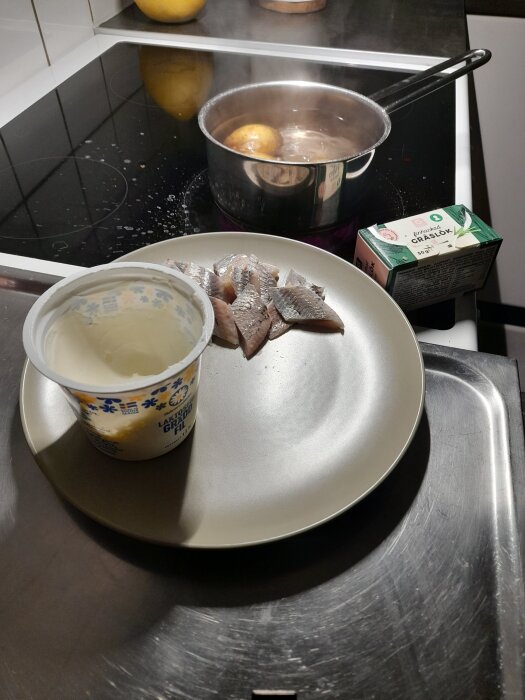 Tallrik med matjessill, en burk gräddfil och paket gräslök. I bakgrunden en kastrull med kokande potatis på spisen.