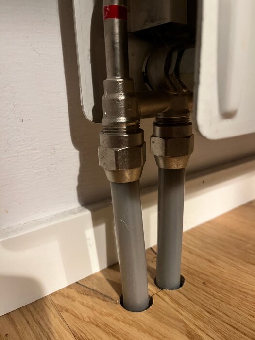 Två gråa rör med kopparrör och kopplingar är anslutna till en väggmonterad radiator, syns mot en vit vägg och trägolv.