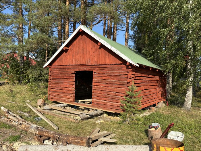 Äldre röd hölada med grönt tak, omgiven av träd och verktyg, som planeras bli en carport.