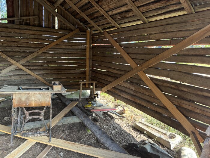 Insidan av gammal hölada med träväggar och diverse byggmaterial på marken, förbereds för omvandling till carport.