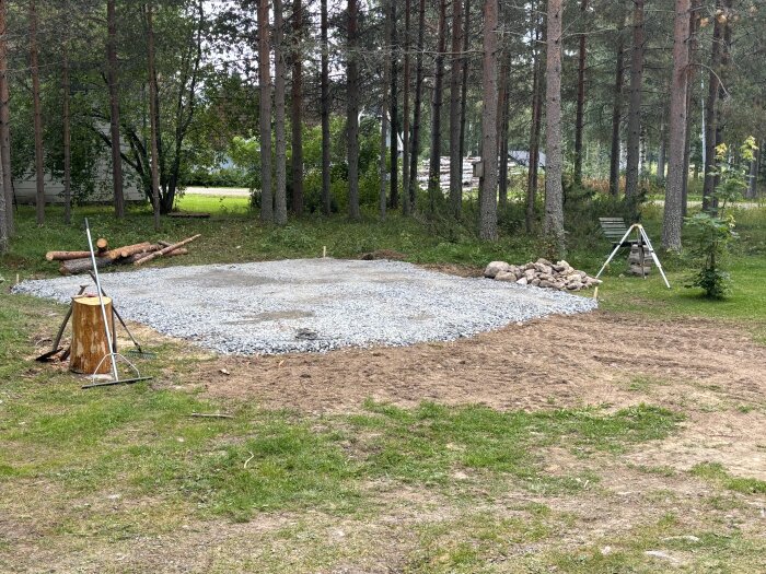 Gruslagd yta i skogsmiljö förbereds för carport, med trädstubbe, spadar och en hög med stenar runt omkring. Träd och buskar i bakgrunden.