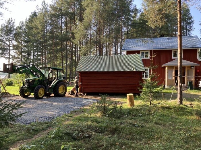 Traktor flyttar en liten röd stuga byggnad vid sidan av ett större rött hus omgiven av träd i en skogsmiljö.