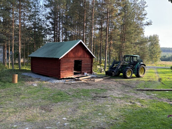 Traktor vid röd timmerstuga i skogsmiljö under byggnation, med grönt grästak och omgivande granar.