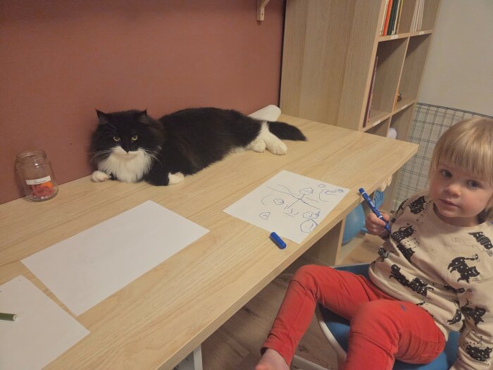 Svartvit katt liggandes på bord bredvid barn som ritar; katt i fokus.