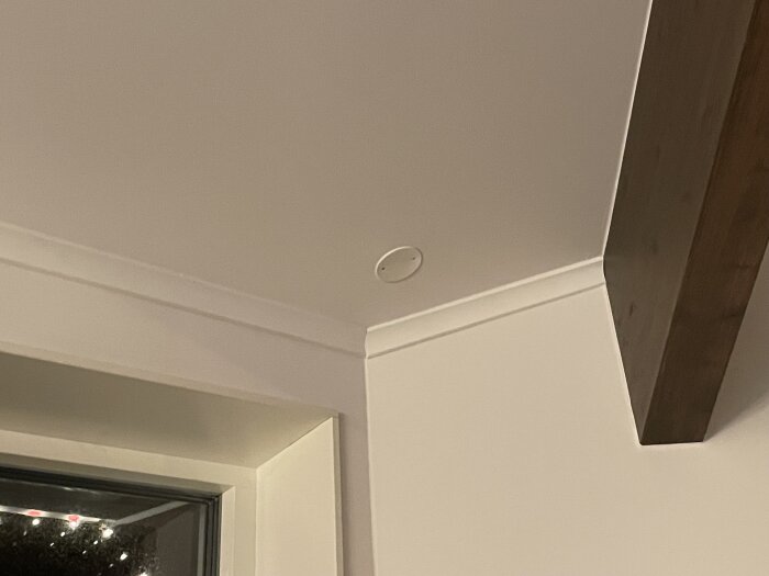 Hörn av ett rum med ett vitt tak och vägg; en rökdetektor syns i taket, en träbalk löper längs väggen, och ett fönster med dekorationer.