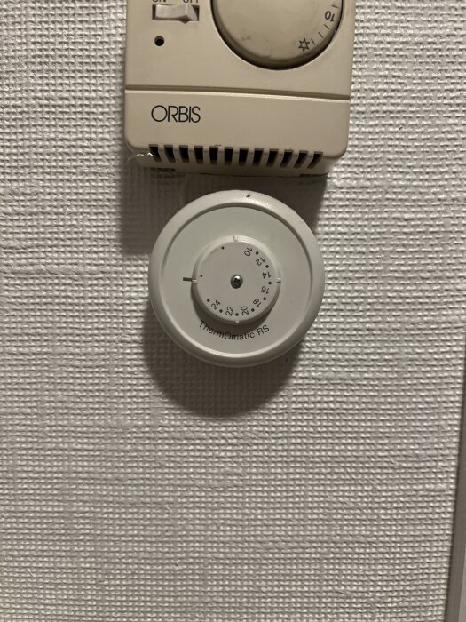 Väggmonterad termostat och en Thermomatic RS-ratt i närbild på en vit strukturtapet.