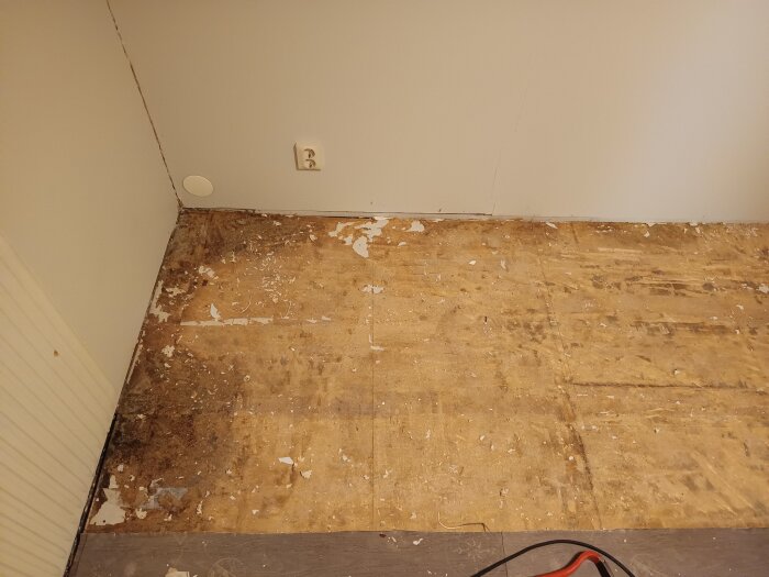 Vattenskadat golv i hörn av rum, plastmatta borttagen, visar blött trä och mörka fläckar nära väggen, spår av fuktskador synliga på underlaget.