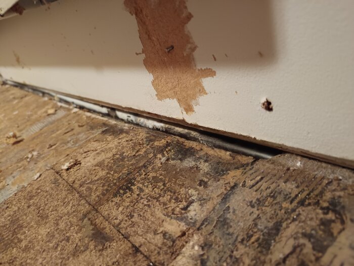 Fuktskadad spånskiva synlig intill en bortbruten väggbit, avslöjar underliggande lager i ett renoveringsprojekt.