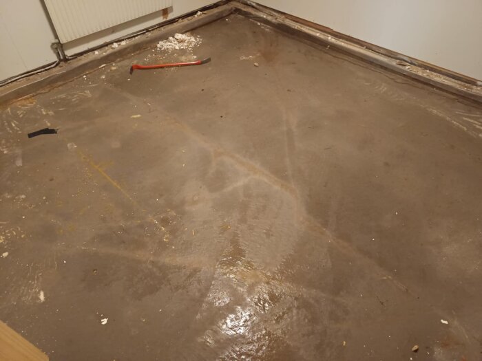 Fuktig betonggolvyta med borttagen plastmatta och spånskiva i ett hörn av ett rum, ett bräckjärn och smutsiga fläckar synliga.