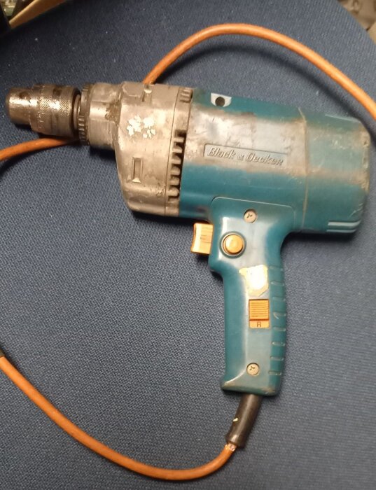 Gammal blå borrmaskin av märket Black & Decker med orange sladd, liggande på en blå yta.