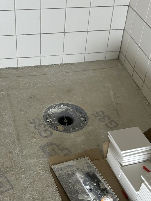 Badrumsgolv under renovering med synlig brunn och tätskikt, kakelplattor och verktyg i förgrunden, brunnsmanschett synlig.