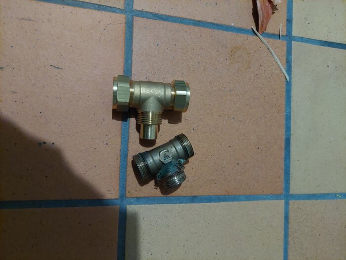 Två T-kopplingar i metall placerade på ett kaklat golv, används för VVS-lösningar i värmesystem.
