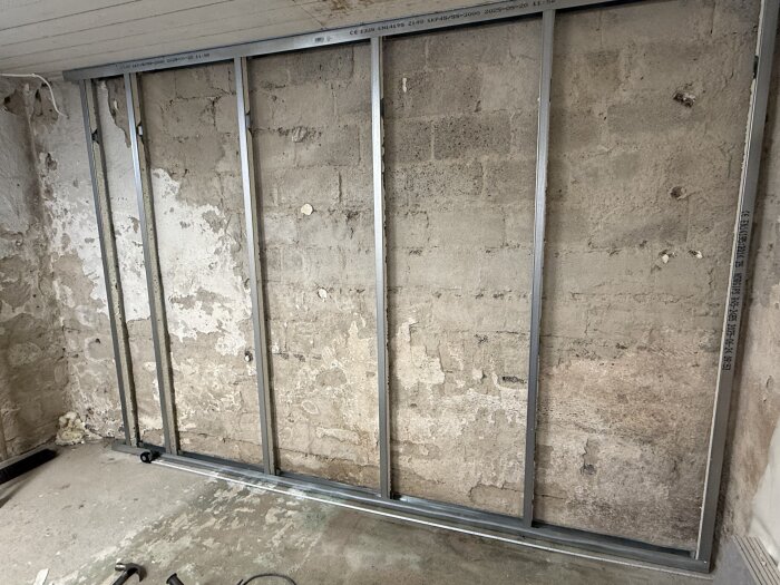 Vägg med synliga stålreglar på en ojämn betongyta, del av ett renoveringsprojekt i ett rum. Verktyg syns på golvet framför väggen.