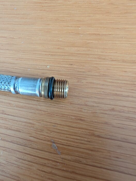 Flexibel slang med gängad koppling och svart o-ring placerad på träbord.