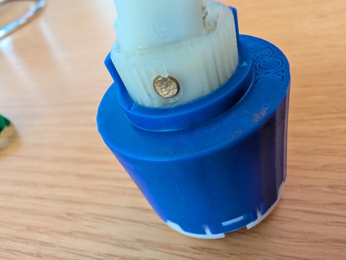 Blå och vit plastdel med skruv och metallklämma, placerad på ett träbord.