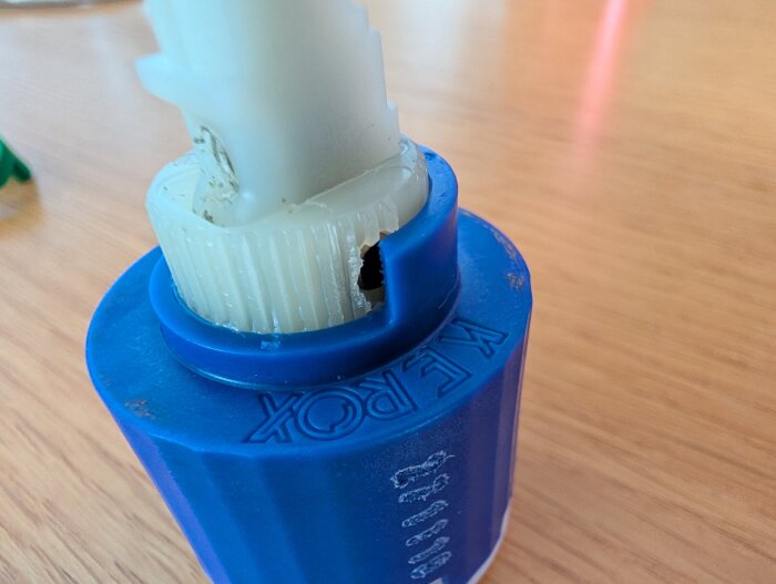 Plastdel med texten "KEROX" och en vit, gängad komponent ovanpå, placerad på ett träbord.