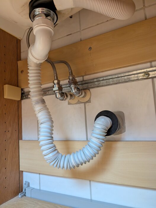 Rör och avloppsanslutningar i ett badrum med exponerade flexibla slangar, monterade nära en vägg med träpanel och kakel.