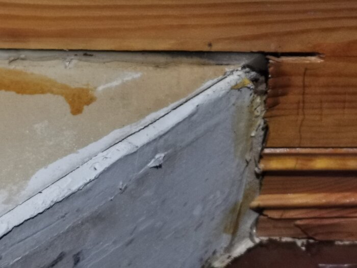 Vindplast syns vid taket där olika material möts, med träpanel och gips. Eventuell vattenfläck på väggen.