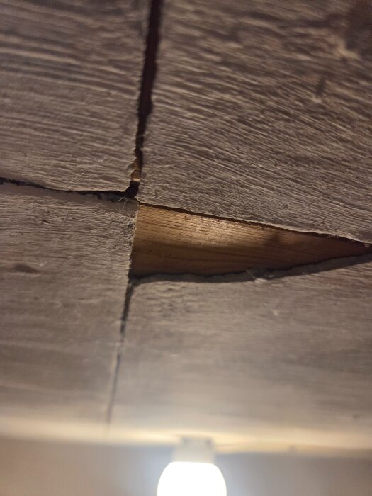Detalj av tak med sprickor och synligt träunderlag, tjocklek cirka 4 mm, i rum med glödlampa.