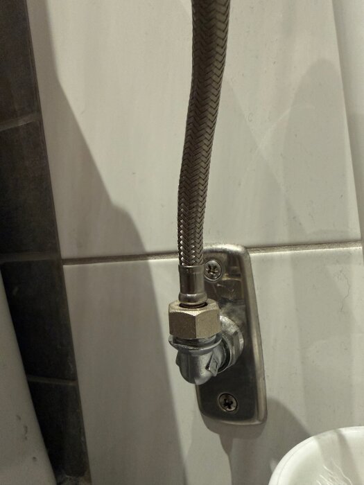Flexibelt anslutningsrör kopplat till vattenuttag på en vit kaklad vägg, relaterat till problem med vattenförsörjning till toalett.
