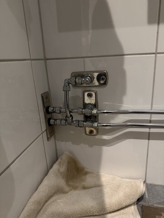 Rörkopplingar och anslutningar i badrum med detalj av vattenanslutningar mot kaklad vägg, beige handduk på golvet nedanför.