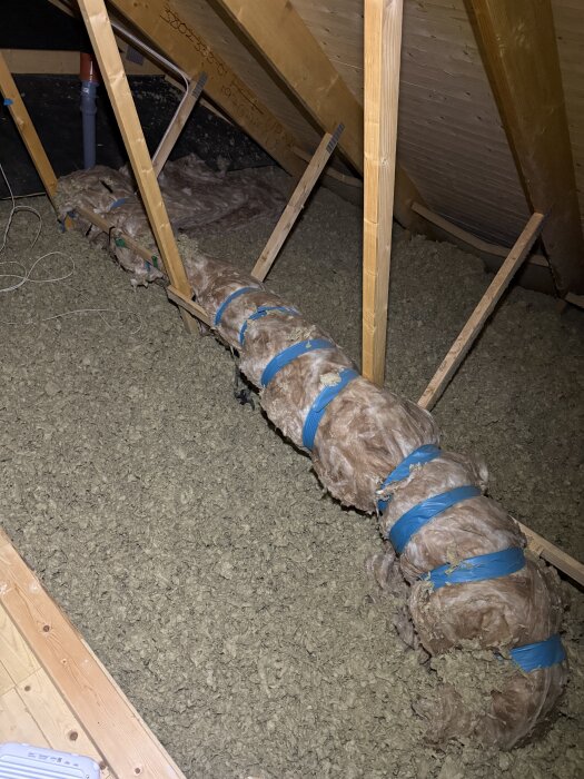 Isolerat rör på en vind, täckt med fiberisolering och blå tejp, synlig trästruktur i taket och paneler på golvet.