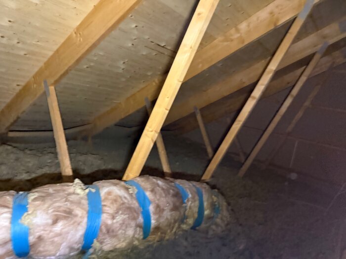 Isolerat rör med blå tejp på vind, omgiven av träbjälkar och isoleringsmaterial.