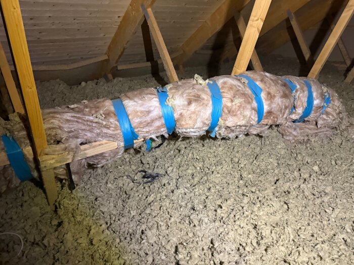 Isolerat rör på en vind sett ovanifrån, med blå tejp håller fast isoleringen. Träbalkar syns i bakgrunden av bilden.
