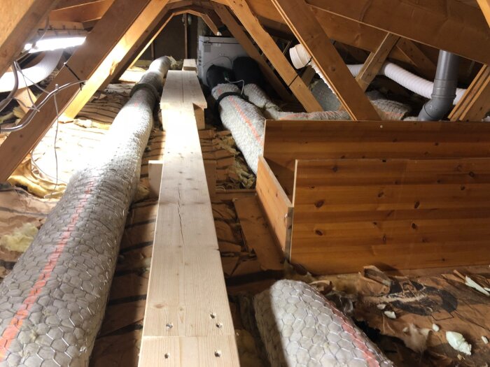 Vindsutrymme med isolerade ventilationsrör omgivna av nät, trästomme synlig ovanför.