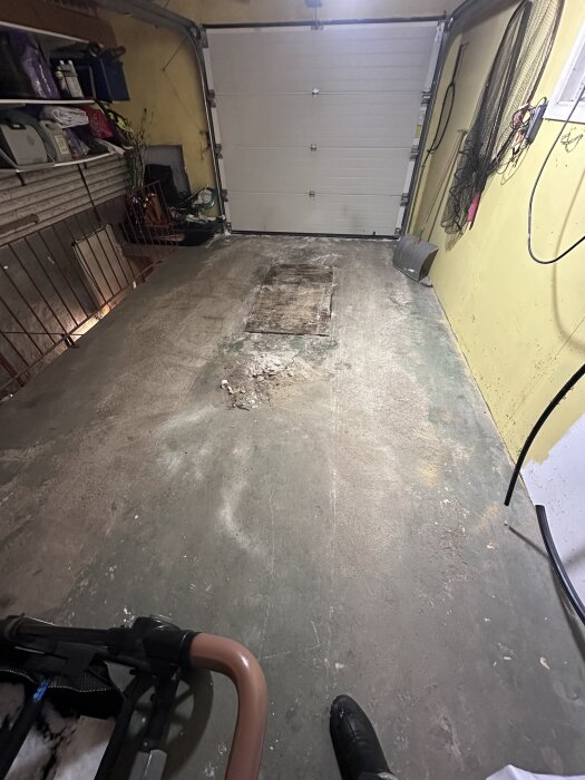 Garagets betonggolv med slitna spår och en platta i mitten, omgivande väggar med förvaring och en stängd garageport i bakgrunden.