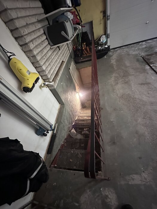 Trappa i garage med metallräcke, sedd uppifrån. Betonggolv, hyllor och förvaringsföremål synliga. En arbetslampa lyser på väggen.