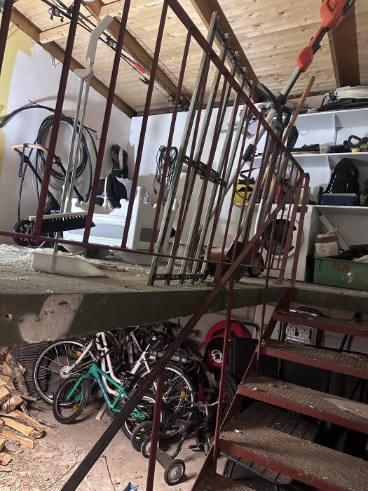 Garage med betonggolv, cyklar och verktyg på hyllor. Trappa och räcke synligt.