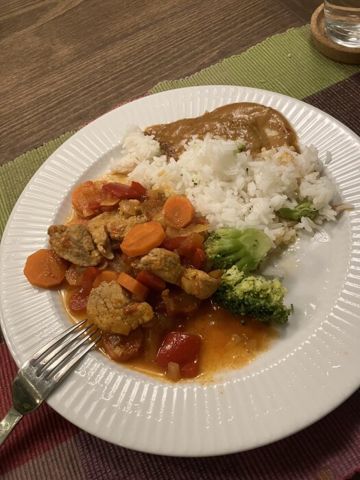 Tallrik med fläskfilégryta, morötter, paprika, ris, broccoli och jordnötssås, serverat på ett bord med grön och lila bordsduk.