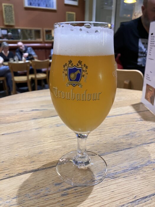 Ett glas öl med skum på en träbord, omgiven av människor i en pubmiljö.