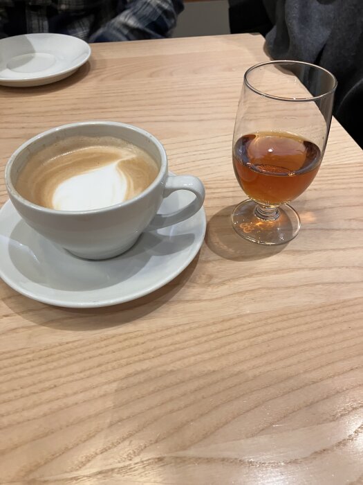 En vit kopp cappuccino på fat bredvid ett glas bärnstensfärgad dryck står på ett träbord.