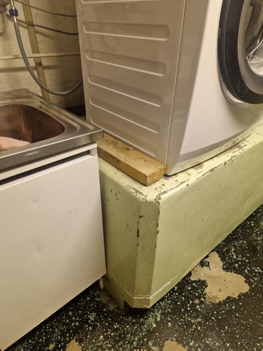 Tvättmaskin på betongfundament i källare med slitet golv. Diskho bredvid och synliga rör bakom. Spår av fukt på golvet.