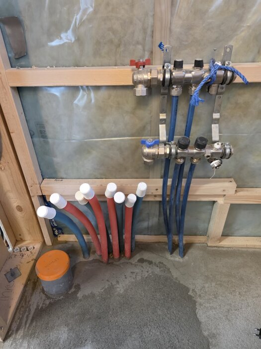 Rörinstallation vid en vägg med flera rör som går upp ur betongplattan, både blå och röda rör syns. Vattenfördelare monterade på trä.