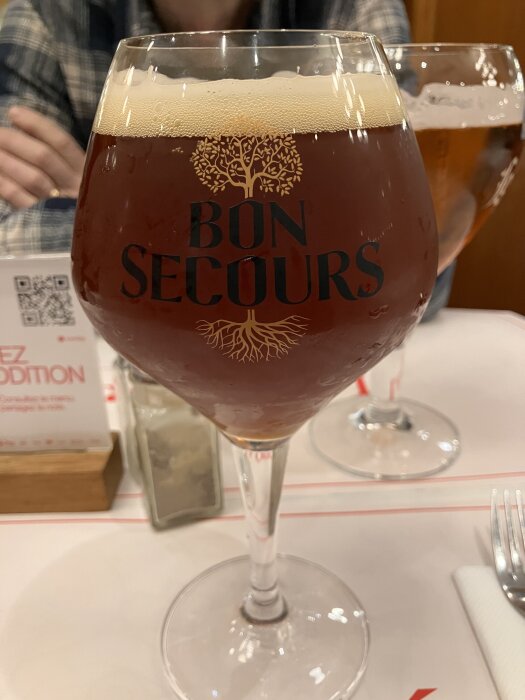 Ett glas med mörk öl märkt "Bon Secours" på ett dukat bord med bestick och en saltströare.