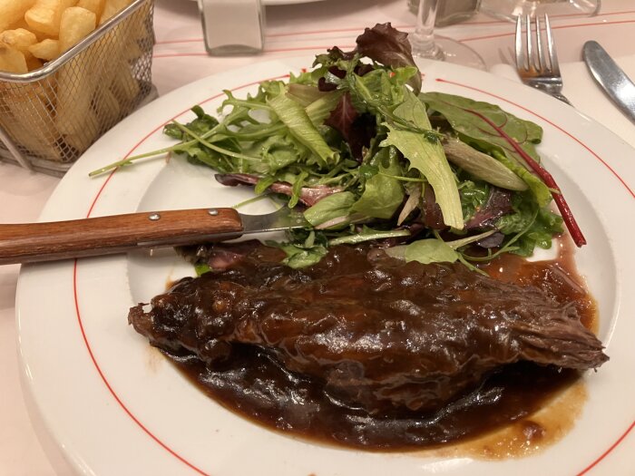 Tallrik med kött i brun sås och en grönsallad. Bredvid finns en liten korg med pommes frites. En kniv ligger på tallriken.
