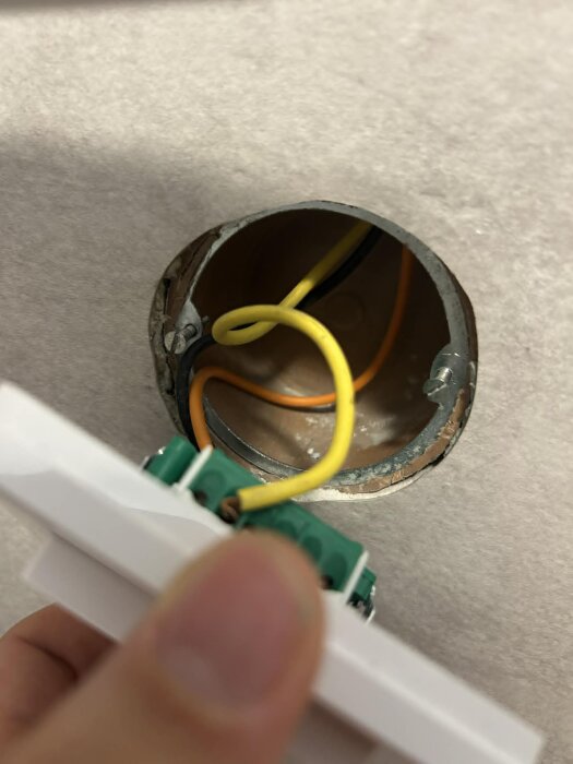 Elektrisk installation med gul och svart kabel i ett uttag, en hand håller en vit plastplatta med gröna anslutningar.