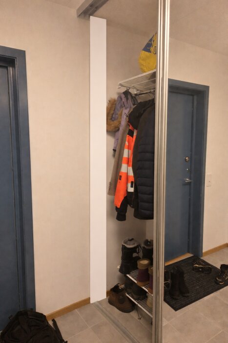 En hall med kläder och jackor hängande bredvid en spegel, hyllor med skor och en blå dörr i bakgrunden.