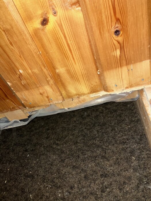 Träpanel möter golv med plastfilm synlig längs kanten.