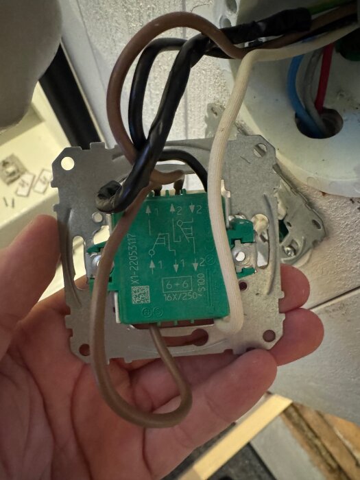 Elektrisk koppling med flera kablar, hålls i hand. En grön komponent med symboler och text visas, del av en fråga om trappkopplingar i ett diskussionsforum om bygg.