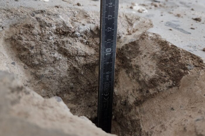 Närbild av mätsticka i betonggolvets hålighet, visar ungefär 10 cm djup, omgivet av ojämna kanter och damm.