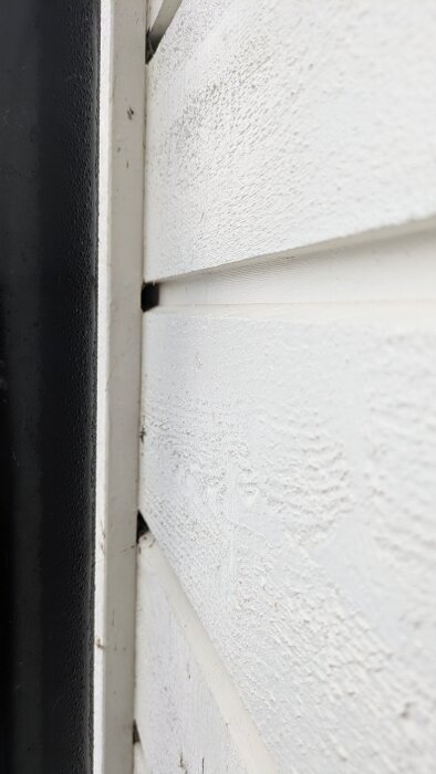 Närbild av vit fasadpanel intill en svart kant. Mellanrummen visar potentiella ventilationshål vid knutbrädan.