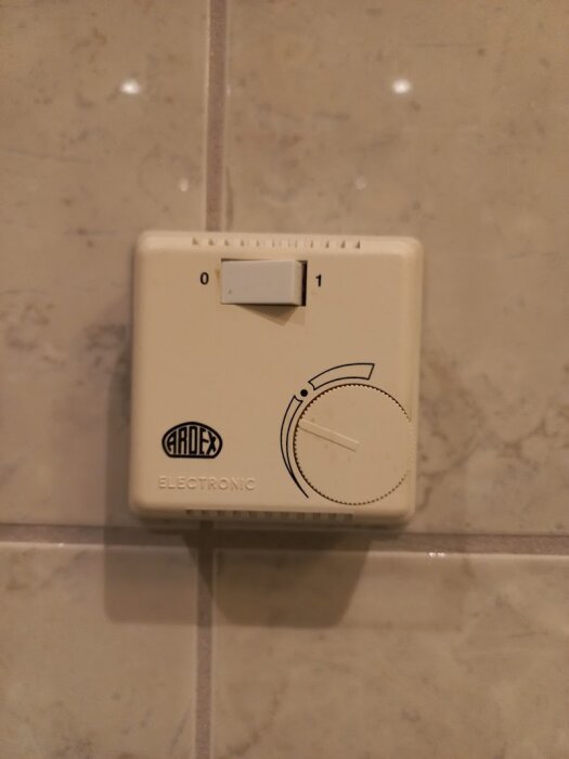 Gammal termostat för golvvärme i badrum, vit med vred och skala markerad 0 till 1, installerad på kakelvägg.