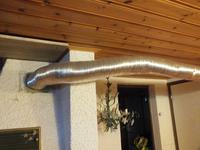 Korrugerad aluminiumrör är monterat under ett tak av trätakplankor i en gillestuga, förankrat nära en vit väggtextur.