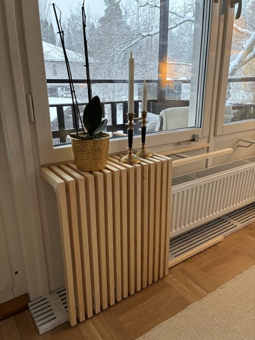 Träradiatorskydd under ett fönster, med en guldfärgad kruka med en växt och två ljusstakar ovanpå, vinterlandskap syns utanför.